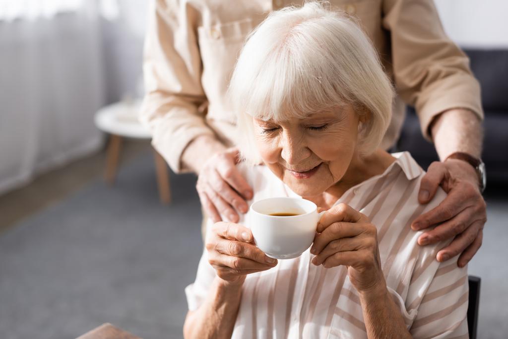 Επιλεκτική εστίαση του ηλικιωμένου άνδρα αγκαλιάζει τη σύζυγο με φλιτζάνι καφέ στο σπίτι  - Φωτογραφία, εικόνα