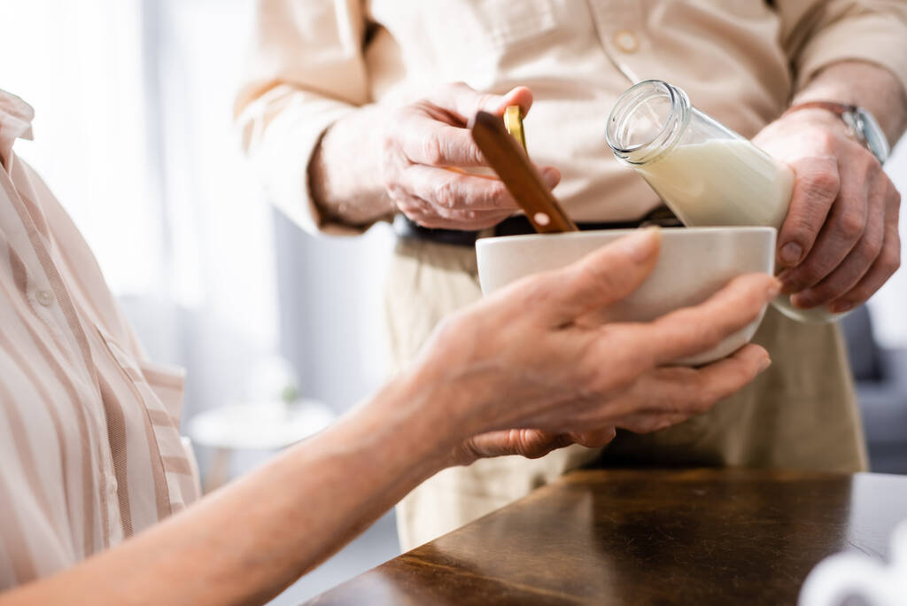 Καλλιεργημένη άποψη του ηλικιωμένου άνδρα ρίχνει γάλα, ενώ η σύζυγος κρατώντας μπολ στο τραπέζι  - Φωτογραφία, εικόνα