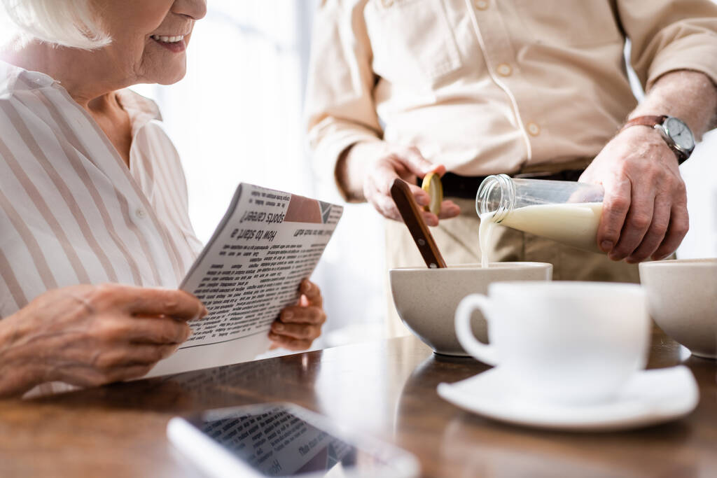 Widok człowieka wylewanie mleka w misce w pobliżu uśmiechniętej żony trzymając gazetę podczas śniadania w kuchni  - Zdjęcie, obraz