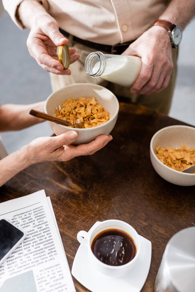 Vágott kilátás nő gazdaság tál gabonafélék, míg a férj öntés tej közelében kávé, újság és okostelefon az asztalon  - Fotó, kép