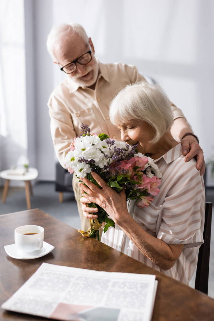 Enfoque selectivo del hombre mayor abrazando esposa con ramo cerca de periódico y taza de café en la mesa
  - Foto, Imagen