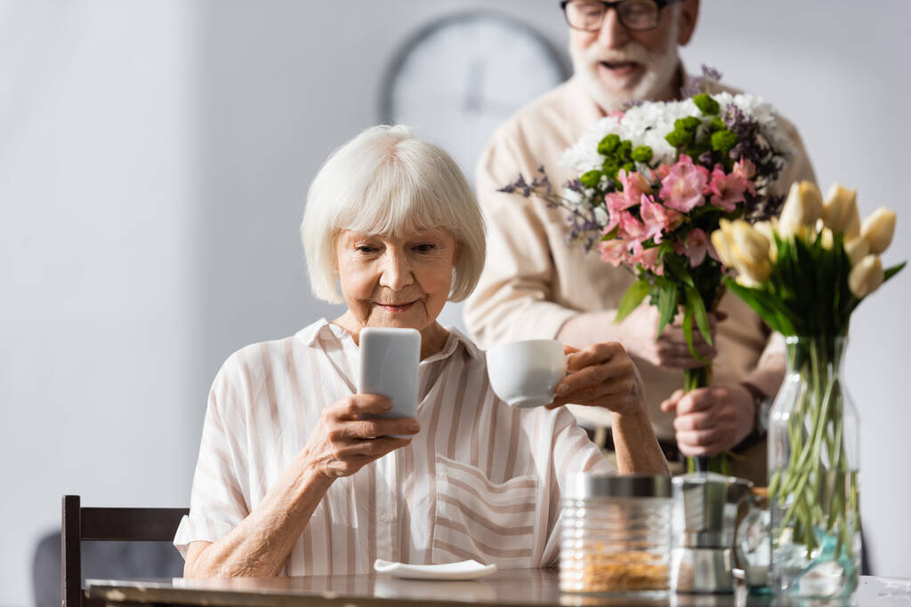 Selektiver Fokus der Seniorin mit Smartphone und Kaffeetasse neben positivem Mann mit Blumenstrauß zu Hause  - Foto, Bild