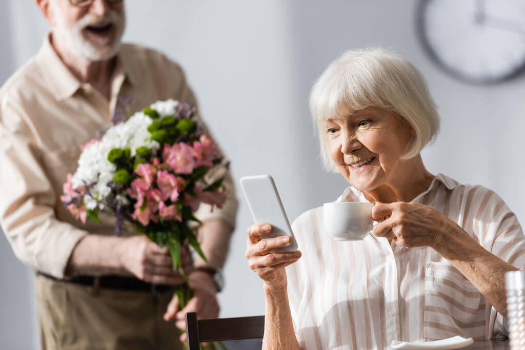 Selective focus of smiling woman holding cup of coffee and using smartphone near husband with bouquet  - Photo, Image