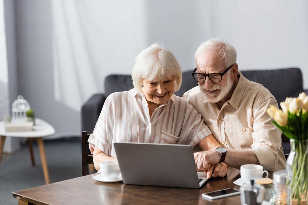 Enfoque selectivo de la sonriente pareja de ancianos utilizando el portátil cerca de tazas de café en la mesa
  - Foto, imagen