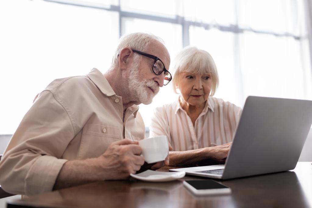 Enfoque selectivo del hombre mayor beber café, mientras que la esposa usando el ordenador portátil cerca del teléfono inteligente en la mesa
  - Foto, imagen