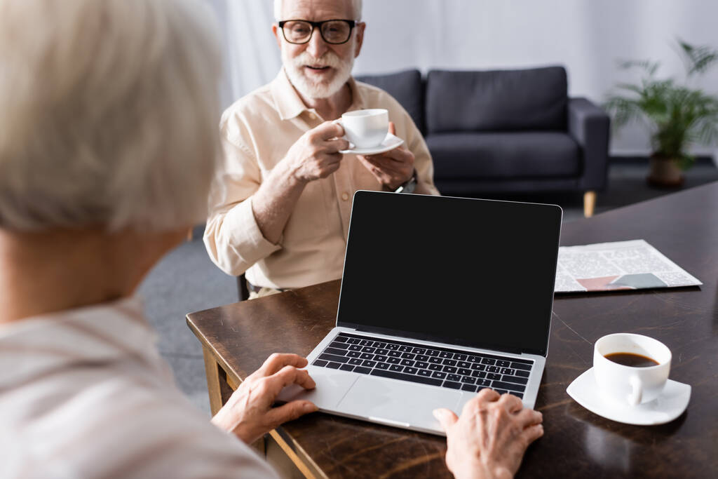 Селективный фокус пожилого мужчины, пьющего кофе рядом с женой с помощью ноутбука за столом
  - Фото, изображение