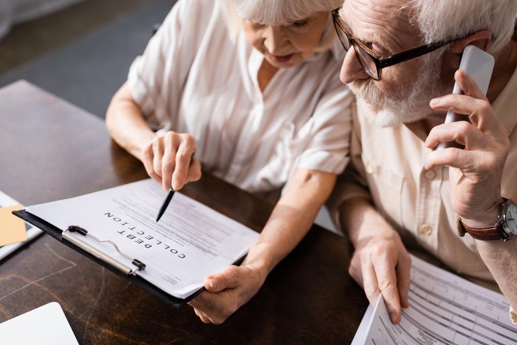 Высокий угол обзора пожилого человека, говорящего на смартфоне рядом с женой, указывающего на документ с надписью о взыскании долгов
 - Фото, изображение