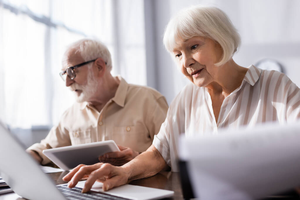 Selektive Fokussierung von Senioren-Paar mit Gadgets in der Nähe von Zeitungen auf dem Tisch zu Hause  - Foto, Bild