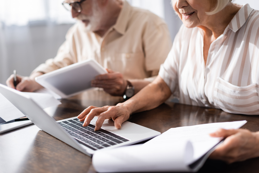 Селективный фокус пожилой женщины, использующей ноутбук и держащей бумаги рядом с мужем с цифровой планшет дома
  - Фото, изображение