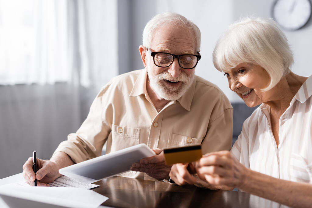 Selektivní zaměření usměvavé starší ženy držící kreditní kartu poblíž manžela s digitálním tabletem psaní na papíře  - Fotografie, Obrázek