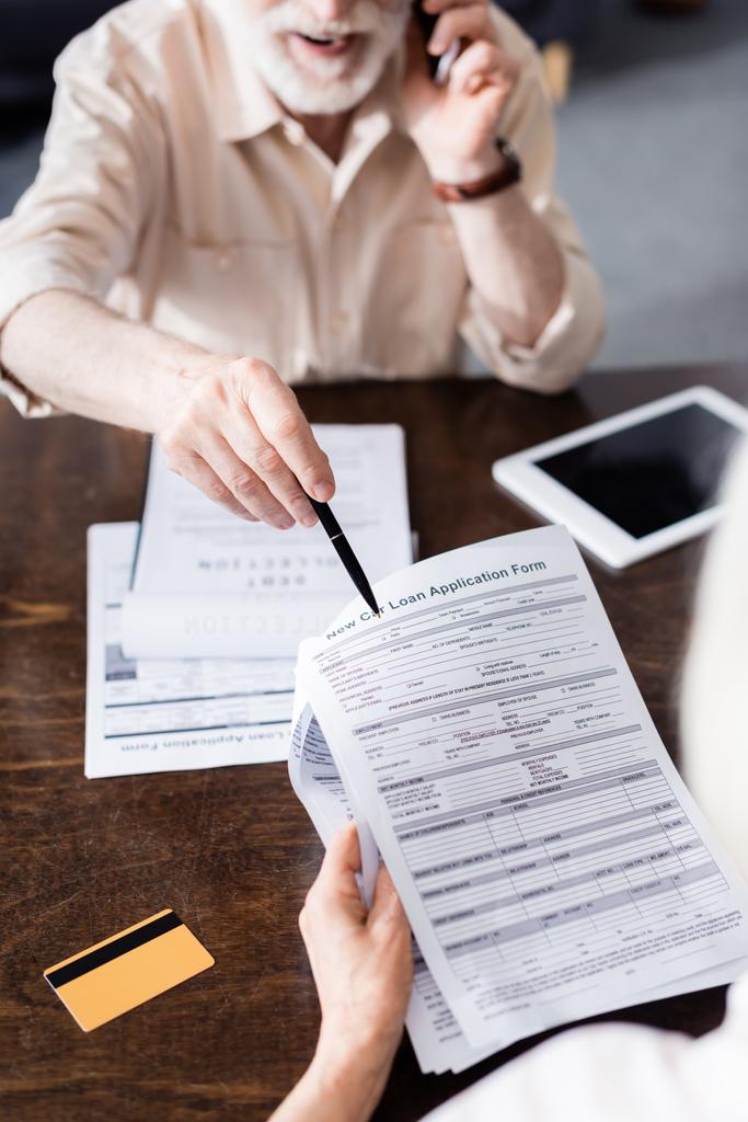 Выборочный фокус человека, говорящего на смартфоне и указывающего на бумаги возле жены и кредитной карты на столе
  - Фото, изображение