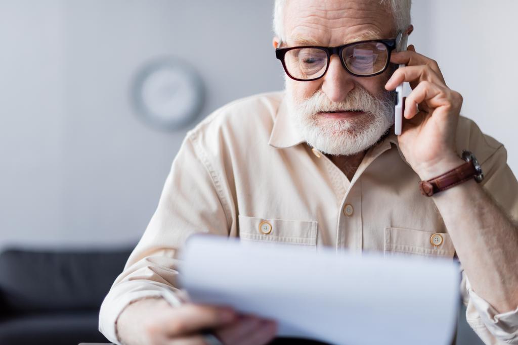 Selectieve focus van senior man holding document en praten op smartphone thuis  - Foto, afbeelding