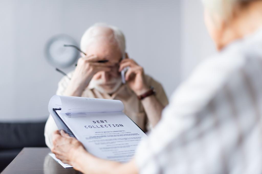 Selective focus of woman holding documents with debt collection lettering while sad husband talking on smartphone  - Photo, Image