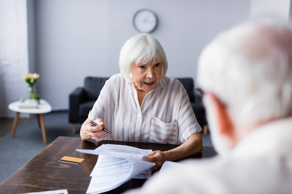 Επιλεκτική εστίαση της ηλικιωμένης γυναίκας που κρατά στυλό και χαρτιά και μιλάει με τον σύζυγο στο σπίτι  - Φωτογραφία, εικόνα