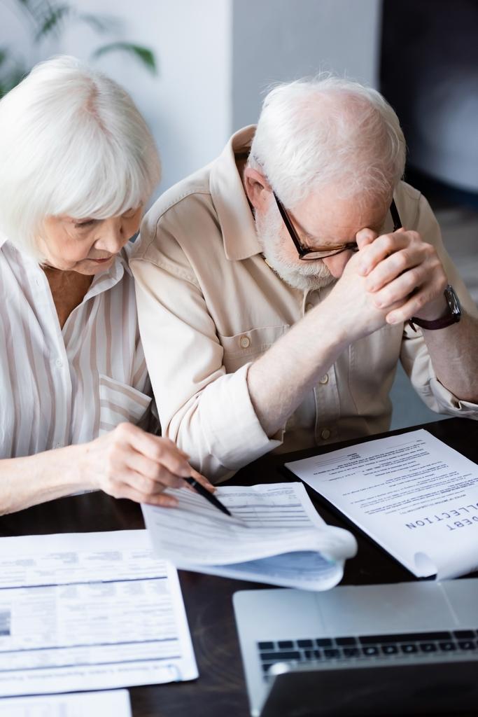 Az idősebb nő szelektív fókusza a szomorú férj közelében lévő dokumentumokra, adósságbehajtási betűkkel ellátott papírra mutatva  - Fotó, kép