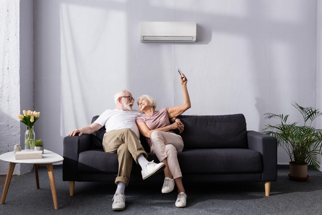 Vrolijk senior paar met behulp van afstandsbediening van airconditioner op de bank  - Foto, afbeelding
