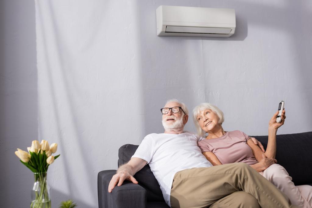 Valikoiva painopiste hymyilevä vanhempi nainen tilalla kaukosäädin ilmastointilaite lähellä aviomies kotona   - Valokuva, kuva
