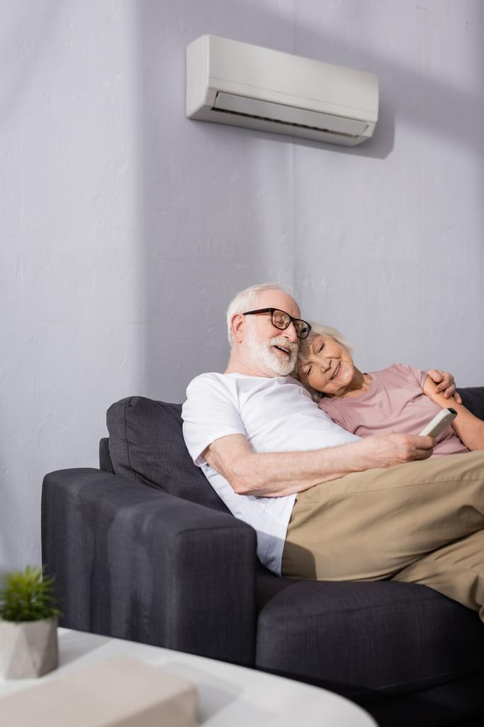 Foco seletivo de sorrir homem idoso segurando controlador remoto de ar condicionado perto da esposa no sofá em casa
  - Foto, Imagem