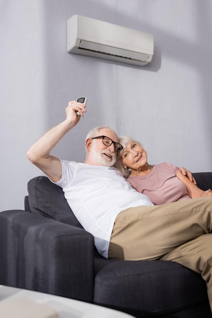 Enfoque selectivo del anciano sonriente abrazando a la esposa y utilizando el controlador remoto de aire acondicionado en el hogar
  - Foto, imagen