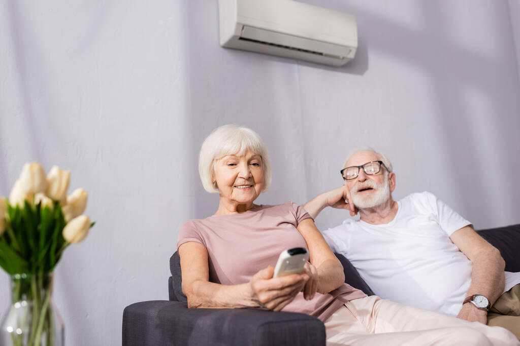 Вибірковий фокус усміхненої старшої жінки, що тримає дистанційний контролер кондиціонера біля чоловіка вдома
  - Фото, зображення