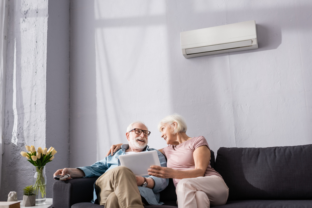 Donna anziana con tablet digitale e guardando sorridente marito con telecomando del condizionatore d'aria a casa  - Foto, immagini
