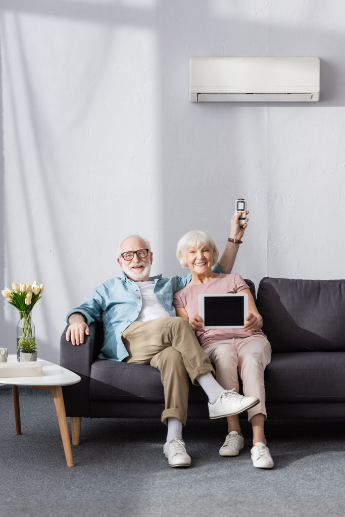 Pareja mayor sonriente mostrando tableta digital y sosteniendo controlador remoto de aire acondicionado en casa
  - Foto, Imagen