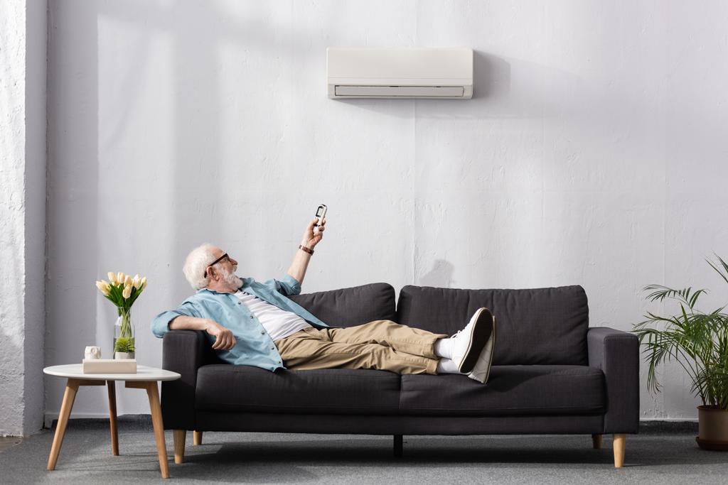 Vidám idős férfi használ távirányító légkondicionáló, miközben ül a kanapén  - Fotó, kép