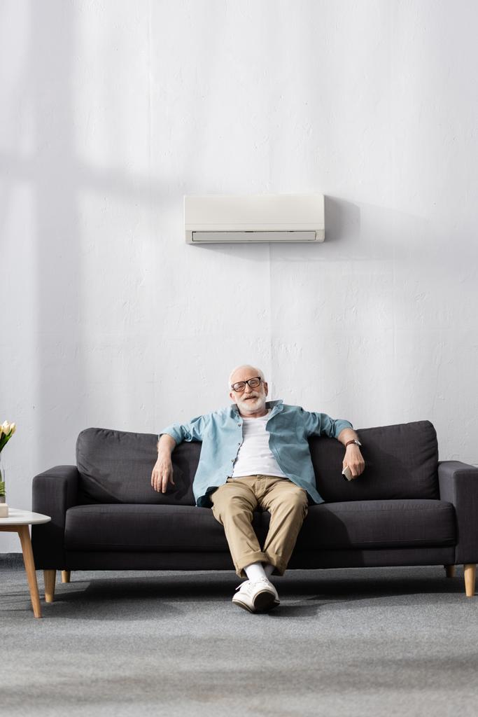 Uomo anziano sorridente che tiene il telecomando mentre è seduto sotto il condizionatore d'aria a casa  - Foto, immagini