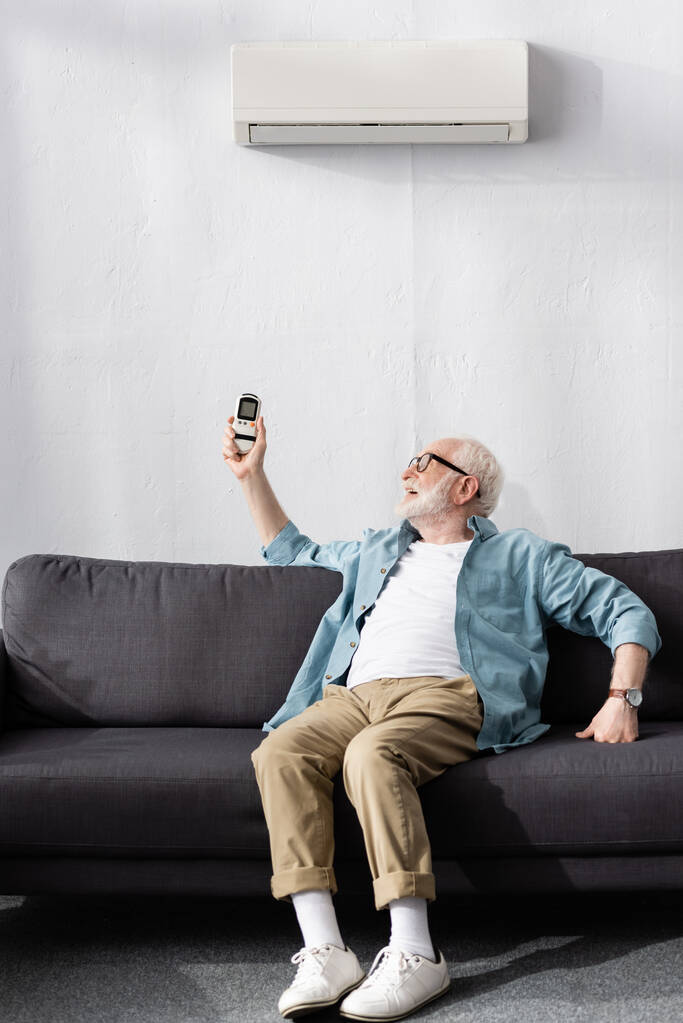 Hombre mayor sosteniendo el mando a distancia mientras está sentado en el sofá bajo el aire acondicionado
  - Foto, Imagen