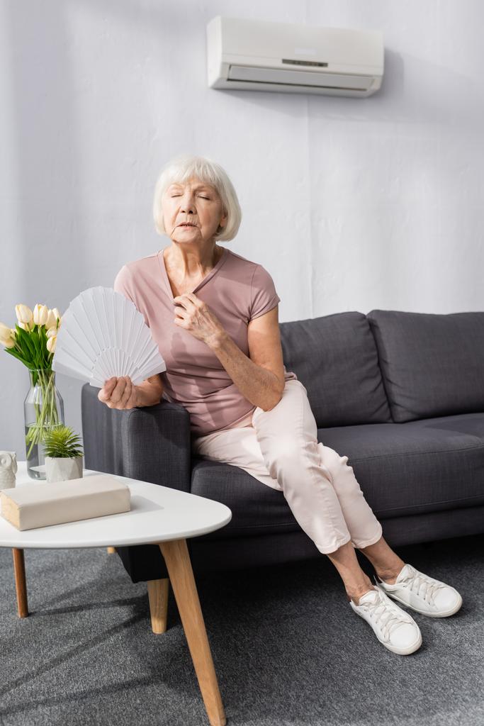 Enfoque selectivo de la mujer anciana sosteniendo ventilador mientras siente calor en la sala de estar
 - Foto, imagen