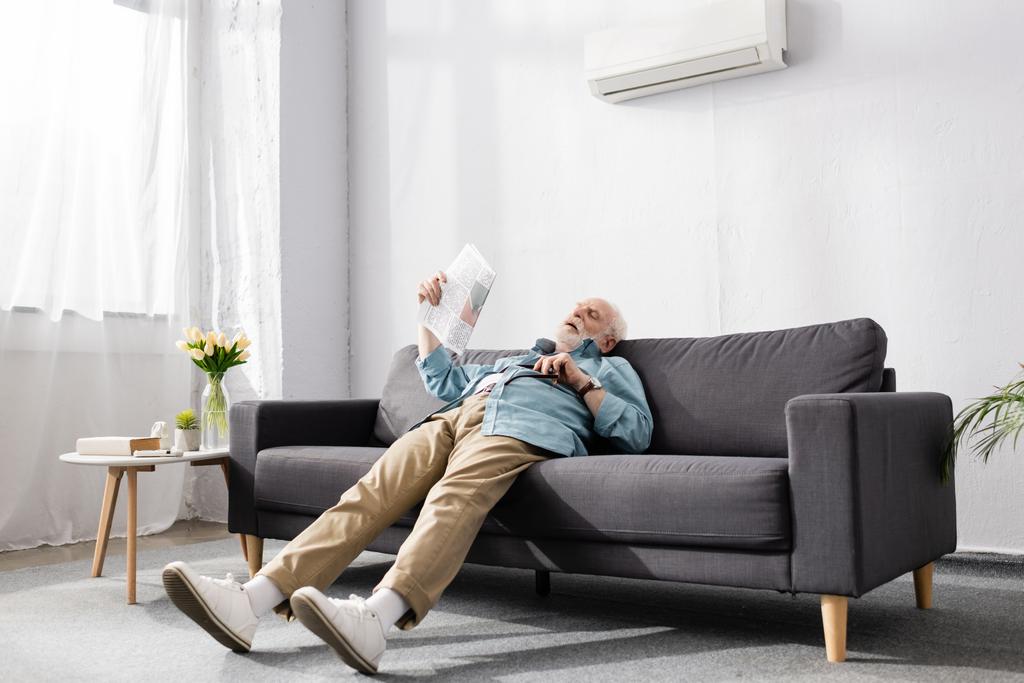 Zmęczony starszy mężczyzna trzymający gazetę na kanapie pod klimatyzatorem w domu  - Zdjęcie, obraz