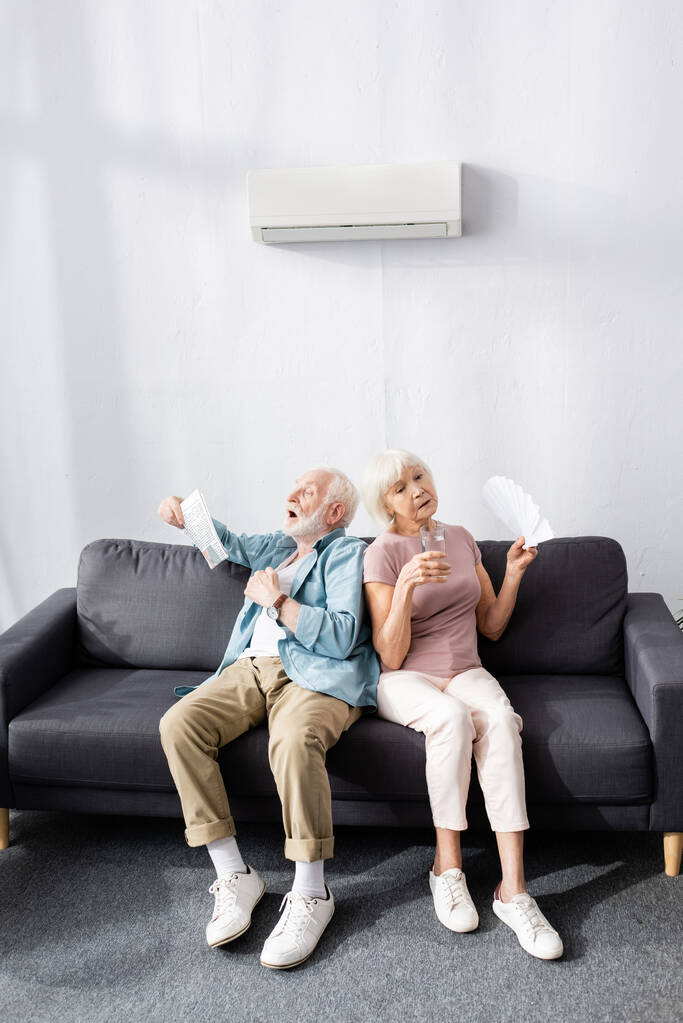 Couple âgé fatigué avec verre d'eau, journal et ventilateur se sentant chaud à la maison  - Photo, image