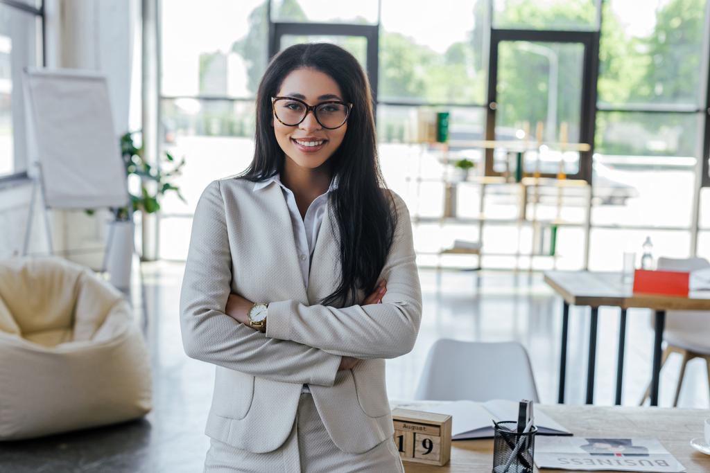 szczęśliwy businesswoman w okularach stojących z skrzyżowanymi ramionami w pobliżu stołu w biurze  - Zdjęcie, obraz
