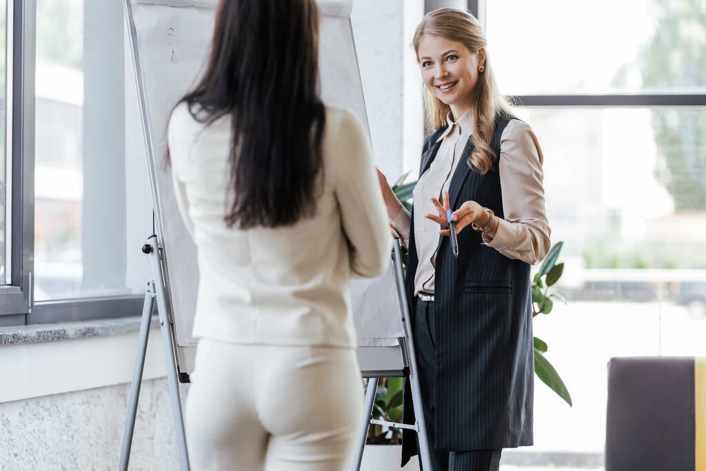 selective focus of happy businesswoman holding marker pen near flipchart and looking at coworker  - Photo, Image