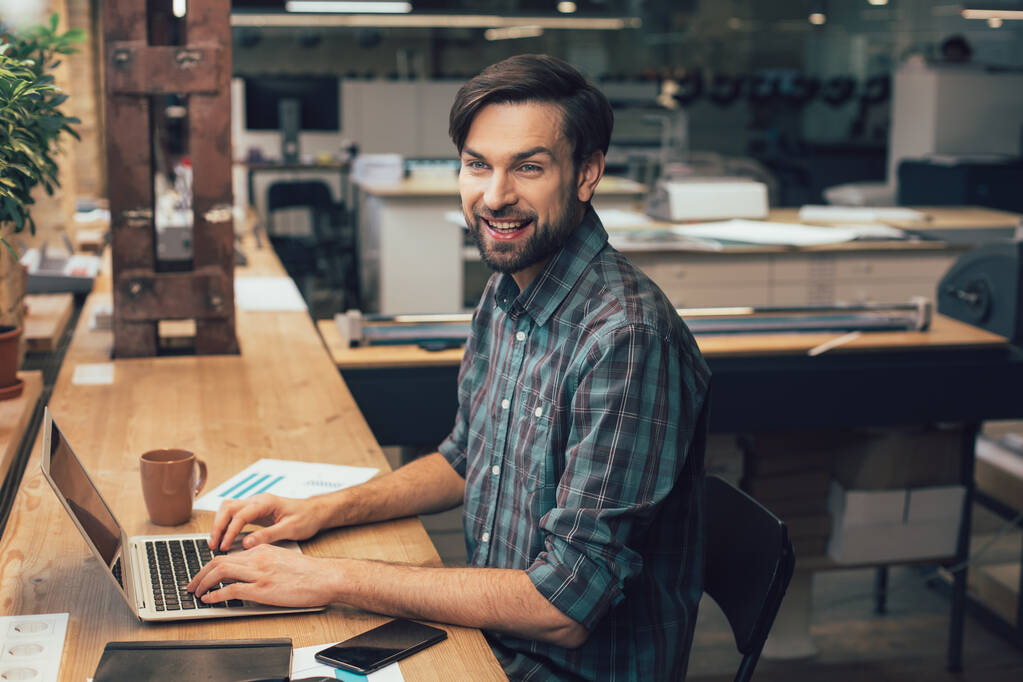 Счастливый работник издательской компании, сидящий за столом с руками на клавиатуре ноутбука и улыбающийся
 - Фото, изображение