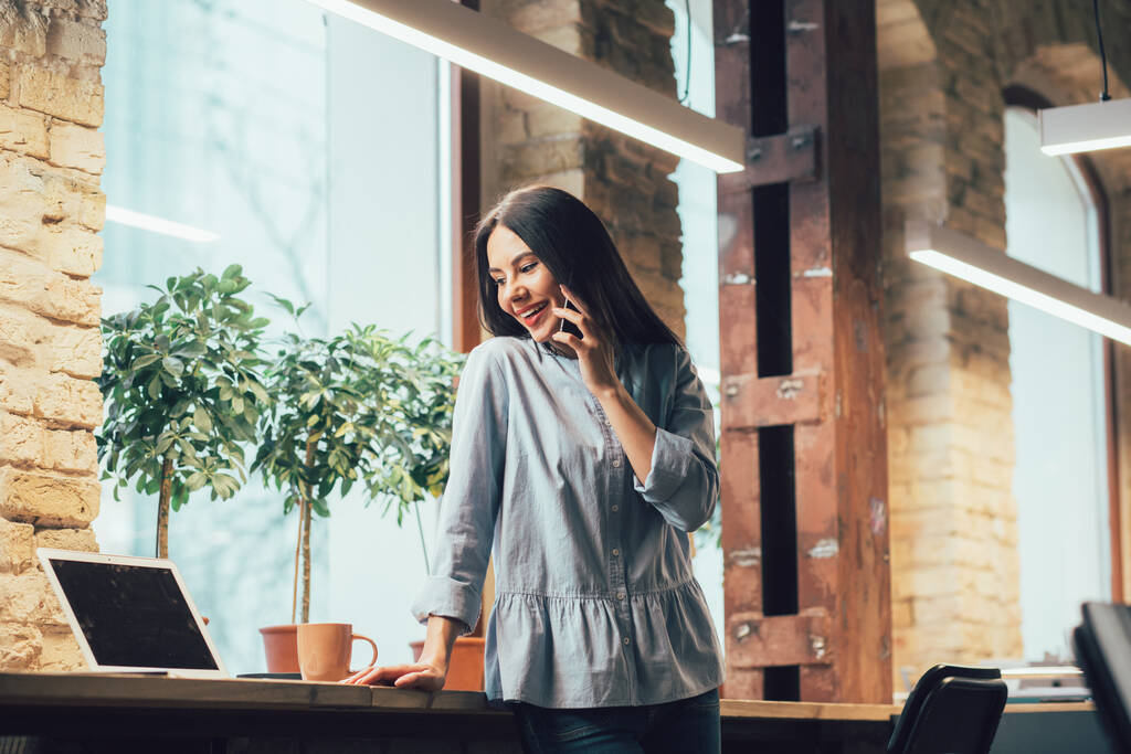 Radosny, piękny pracownik biurowy stojący przy parapecie z kubkiem i laptopem przy jej boku i rozmawiający przez telefon - Zdjęcie, obraz