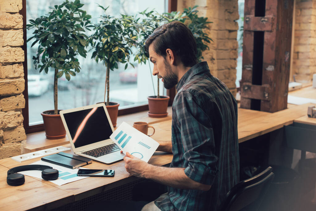 Концентрированный молодой сотрудник смотрит на экран ноутбука и держит графику на листе бумаги. Наушники и ноутбук рядом с ним
 - Фото, изображение