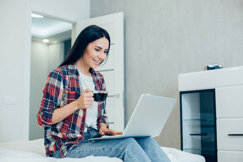 Positiv schöne Frau sitzt auf dem Bett in legerer Kleidung und hält eine Tasse Kaffee, während sie auf den Laptop-Bildschirm schaut - Foto, Bild