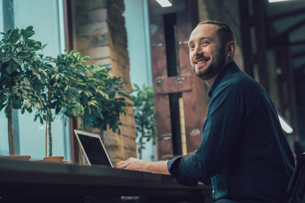 Iloinen komea nuori mies istuu moderni kannettava tietokone toimistossa ja katselee pois hymyillen - Valokuva, kuva