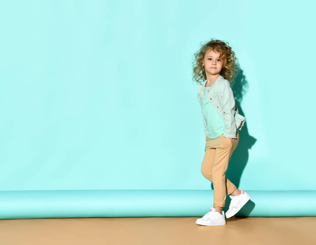 Pretty curly little girl standing near the wall with hands in pockets and calm serious face. Full length portrait on turquoise background - Photo, Image