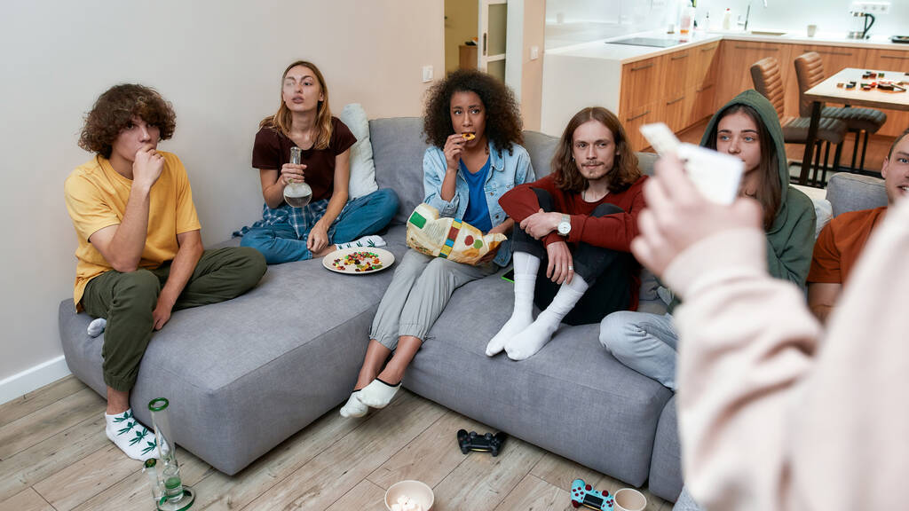Молодые мультикультурные люди едят закуски, играют в видеоигры после курения марихуаны из бонга, сидя на диване. Вид сбоку
 - Фото, изображение