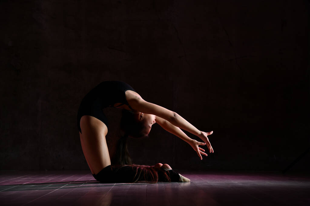 Młoda gimnastyczka w czarnym ciele sportowym i specjalnym obuwiu tworząc gimnastyczną pozę na ciemnym tle - Zdjęcie, obraz