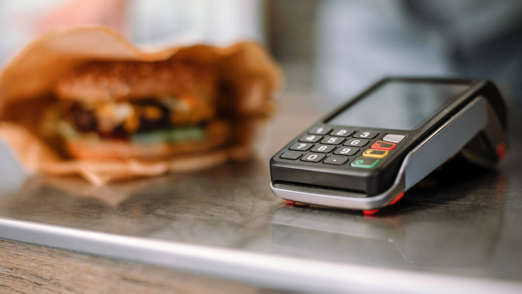 Nahaufnahme eines modernen kontaktlosen Zahlungsterminals mit leckerem Burger im Hintergrund. Street Food Truck Konzept. - Foto, Bild