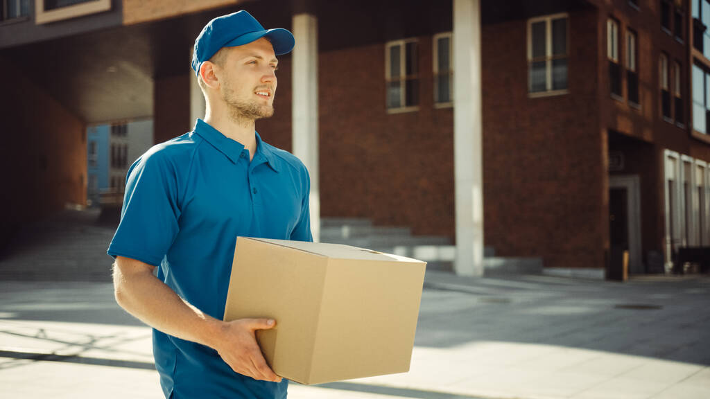 Handsome Delivery Man tient boîte en carton paquet marche à travers le quartier des affaires élégant moderne. Courrier En route pour livrer un colis postal à un client. - Photo, image