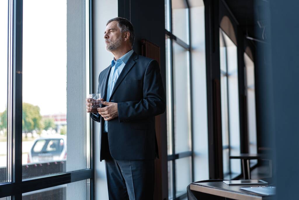 Hombre de negocios maduro bebiendo agua y mirando por una ventana a la ciudad desde un edificio de oficinas
. - Foto, Imagen