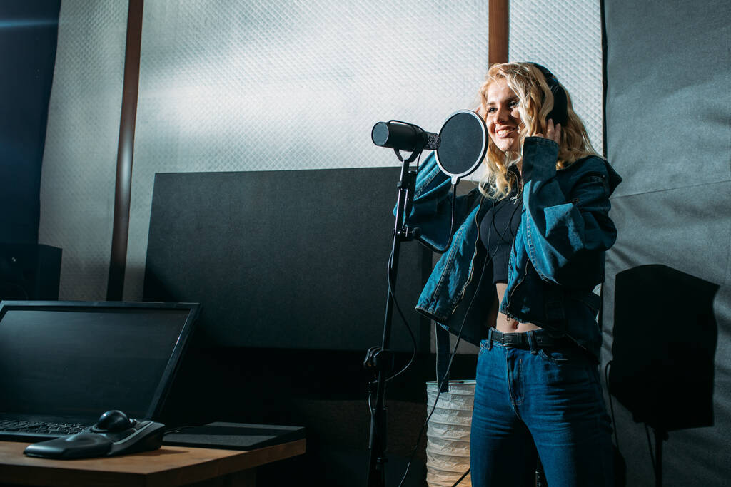 молода красива дівчина, блондинка, співачка, робить запис в студії звукозапису, вона стоїть поруч з мікрофоном на темному тлі, у неї гарна посмішка і емоції. звукорежисер сидить на комп'ютері і контролює процес
 - Фото, зображення