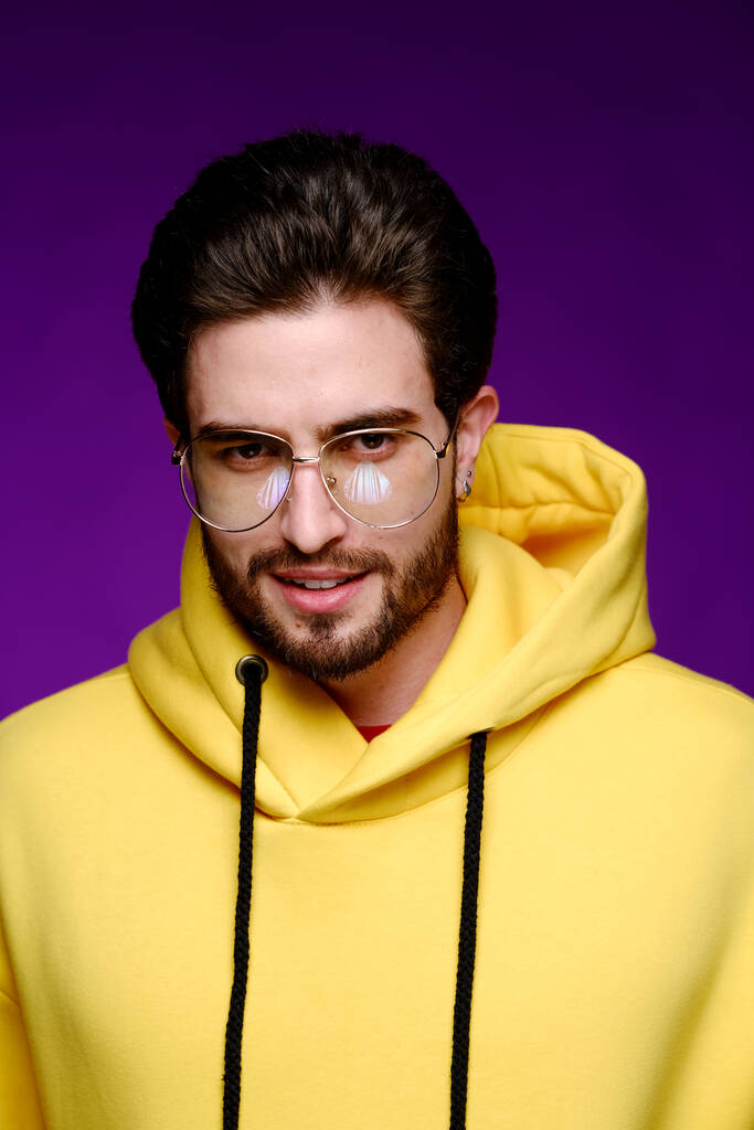 眼鏡で25-30年の若い男と黄色のスウェットシャツは紫の背景に感情的にポーズ.  - 写真・画像