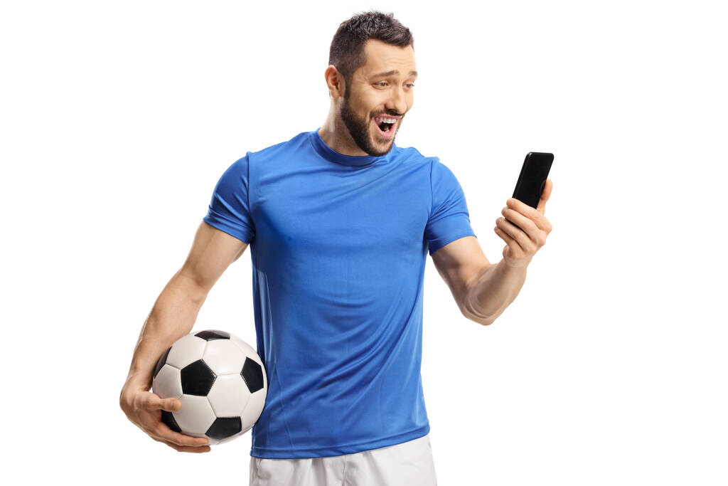 Zaskoczony piłkarz trzymając piłkę i patrząc na telefon komórkowy odizolowany na białym tle - Zdjęcie, obraz