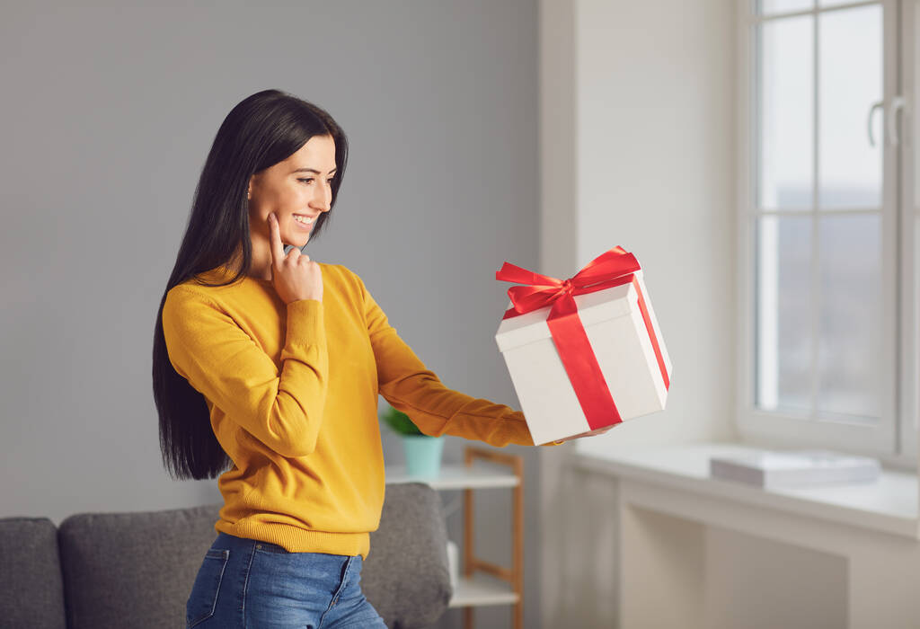 Щаслива дівчина з подарунковою коробкою з червоною стрічкою, що сидить у кімнаті
. - Фото, зображення