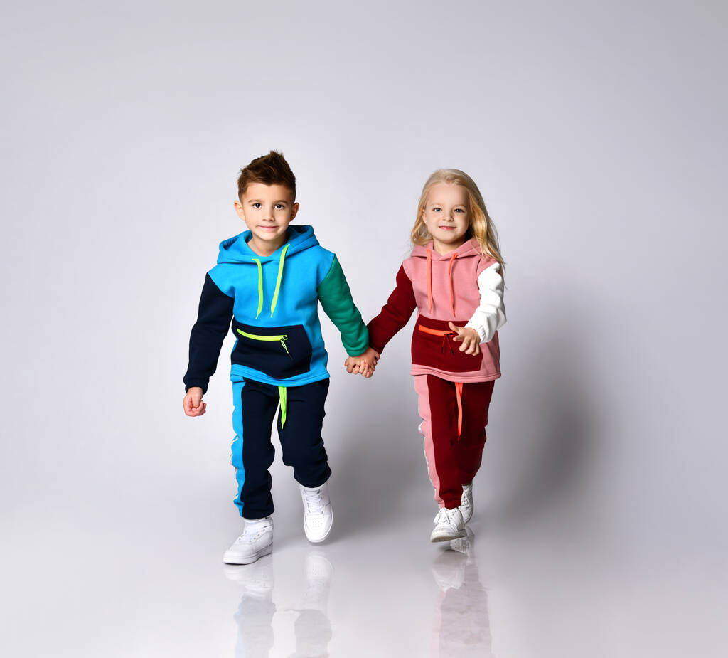 Crianças, menino e menina, em ternos esportivos coloridos e tênis. Eles estão de mãos dadas, correndo, isolados no fundo do estúdio branco
 - Foto, Imagem